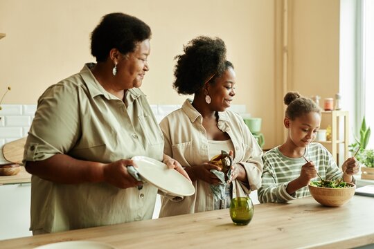Side view of smiling African American mother and granny wiping dishes at large wooden counter watching little girl making fresh salad at cozy beige kitchen
