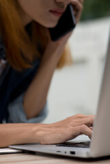 Fototapeta premium Working Woman multitasking on phone and laptop. Close-up of asian busy businesswoman using computer while talking on smartphone, showcasing multitasking and modern communication-vertical image.