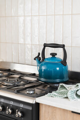 Bright blue kettle on a gas stove, black free standing oven, vertical textured wall tile