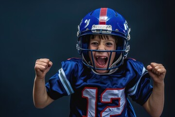 Young boy wears blue football jersey and matching helmet, mouth open in excitement as he celebrates on dark background. Intense moment captured in high-end studio photo.