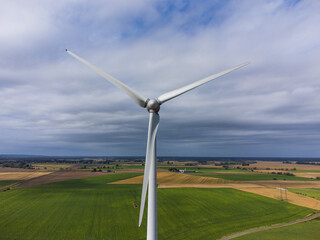 Fototapeta premium Aerial front view of a wind turbine in the wind. Sweden, summer, green and yellow fields and farmland. Cloudy with sunrays illuminating the wind turbine.