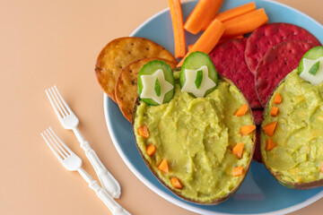 Snack for kids including creamy avocado in the form of crocodile, vegetables crackers on ceramic plate over beige background.