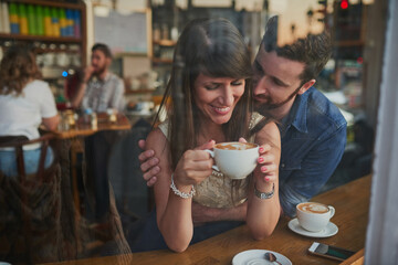 Coffee shop, happy and couple hug in window with drink, caffeine beverage and cappuccino in cafe. Dating, love and man and woman relax in restaurant for bonding, relationship and romance on weekend