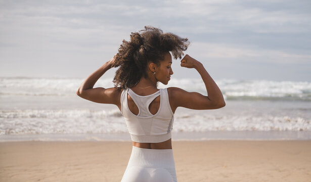 Back view of fit black woman flexing arms towards the sea at the beach. Fitness motivation and success concept. Successful sporty female exercising outdoor.