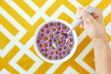 Bowl, spoon and hands with flowers for eating cereal, porridge or breakfast meal by abstract table. Food, plants and high angle of person with floral for nourishment, nutrition or vitamins of nature.