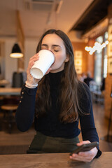 Beautiful young woman in cafe drinking coffee holding cell phone