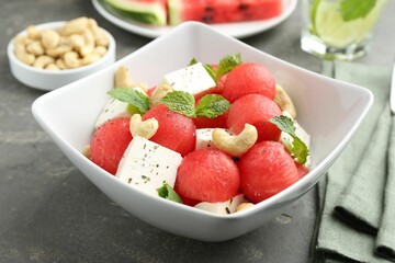 Delicious watermelon salad with feta cheese, cashew nuts and spices in bowl on grey textured table, closeup