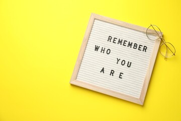 Letter board with phrase Remember Who You Are and glasses on yellow background, top view. Space for...