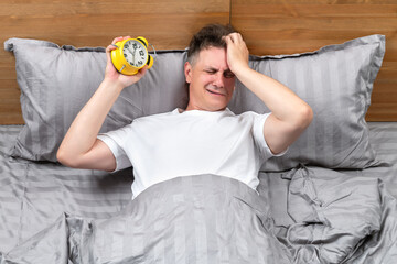 Upset man lying in bed holding yellow alarm clock and facepalming. Concept of overslept for work