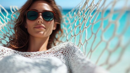 A woman relaxes in a hammock by the ocean, wearing dark shades and a crochet top. The scene reflects tranquility and carefree relaxation under the warm sunlight.