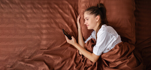 Hispanic woman lies in bed with smartphone, top view. Online call to husband in bedroom. Modern technology