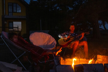 The family is sitting around the campfire, man plays the guitar, a woman listens and sings along, my daughter wrapped herself in a blanket. Family summer evening in the courtyard of the house