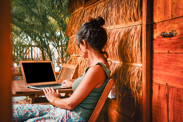 Back view caucasian hipster woman sit by computer outside eco-friendly straw house building guesthouse. Freelance work in south-east asia remotely isolated in tropical nature concept © Evaldas