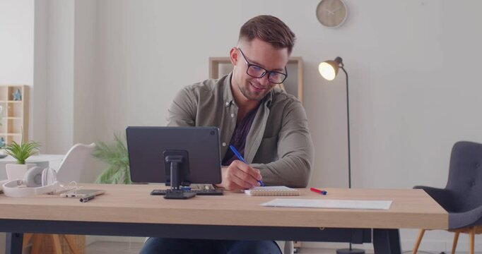 Happy man studying online. Young student in eyeglasses sitting at desk at home, using tablet computer, typing on mini keyboard, enjoying educational research course, learning interesting information