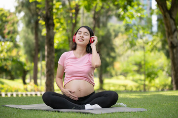 Fototapeta premium Young Asian pregnant woman enjoys listening to music with headphones in the park