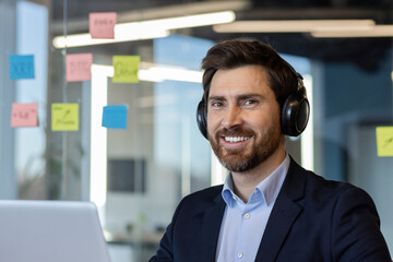 Smiling businessman wearing headphones works at office desk. Focused on computer tasks, surrounded by colorful sticky notes. Represents modern workplace, productivity, and professional concentration