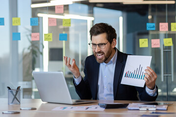 Businessman in modern office showing anger during video call. Holding chart with graph depicting declining sales. Office environment includes laptop, scattered documents