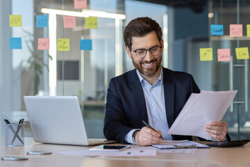 Businessman with glasses and beard sits at office desk, reviewing documents with pen in hand. Surrounded by laptop, notes on window, displaying sense of productivity and focus.