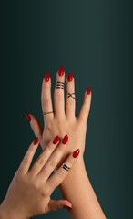 Female hands relaxed, long nails with red gel polish on an emerald green background. A woman with a classic stylish manicure and silver ring set