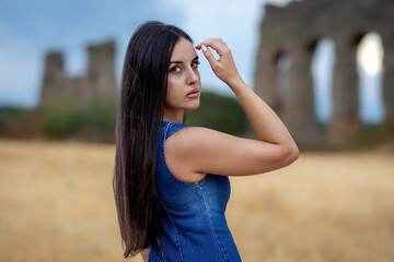 Portrait of brunette girl with long hair and denim dress, in the park of the ancient Roman aqueducts.