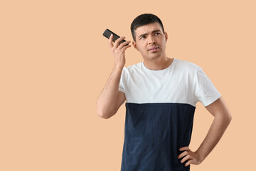 Displeased young man with smartphone listening to voice message on beige background