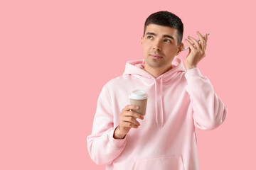 Young man with coffee and smartphone listening to voice message on pink background