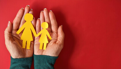 Hands holding a family recreated in yellow paper, consisting of a mother and daughter, on a red background. The person holding the concept of family unity is wearing a green sweater