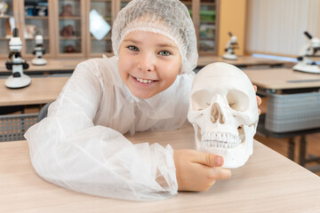 A happy little teenage girl holds an anatomical model of a human skull in her hands. Schoolgirl in a white coat at a science lesson