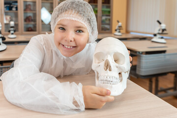 A happy little teenage girl holds an anatomical model of a human skull in her hands. Schoolgirl in a white coat at a science lesson