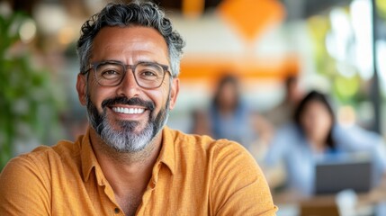 Obraz premium A man with glasses and a gray beard smiles warmly in an inviting cafe, wearing an orange shirt, with a vibrant background that adds to the modern, friendly atmosphere.
