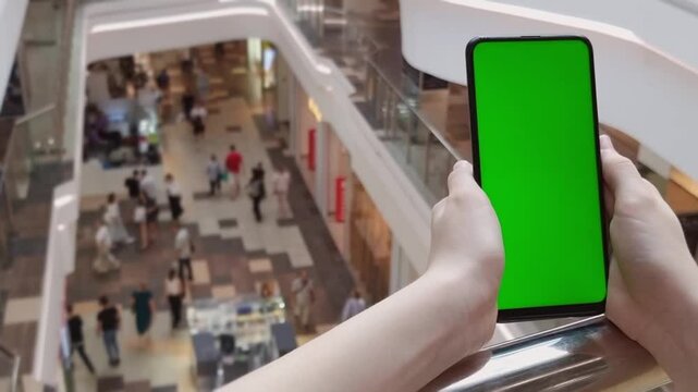The hands of a young girll holds a phone with a green screen in her hand and clicks on the screen, in the background there are people walking with purchases on different floors. . Using a smartphone