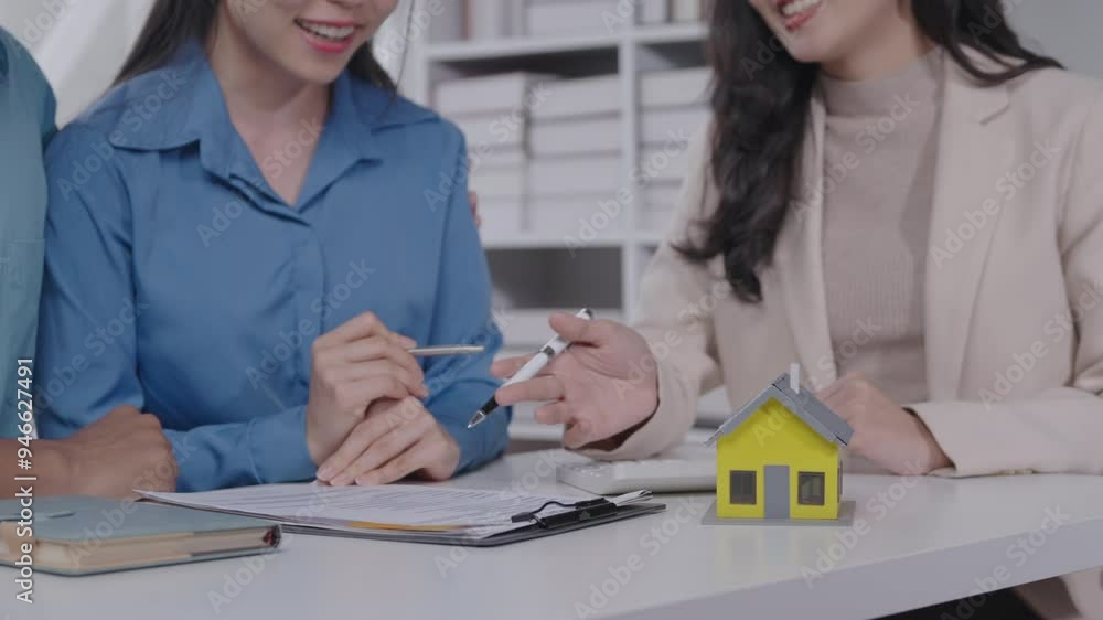 Poster real estate agents sit at a table with a couple, discussing property details while using a small hou