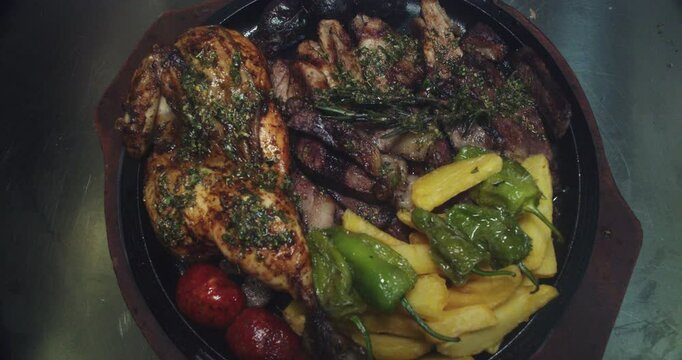 Beautiful slow motion shot of a great grill plate with beef and pork meats, chorizo, potatoes, padron peppers and chimichurri in a restaurant kitchen.