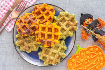 Orange and green waffles on purple plate for Halloween kid's breakfast. Pumpkin waffles and chard waffles over table with Halloween decorations.