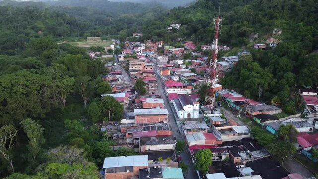 An aerial shot capturing a quaint Venezuelan rural village, surrounded by lush forests, with a cell phone tower standing tall in the center, contrasting modernity with tradition.