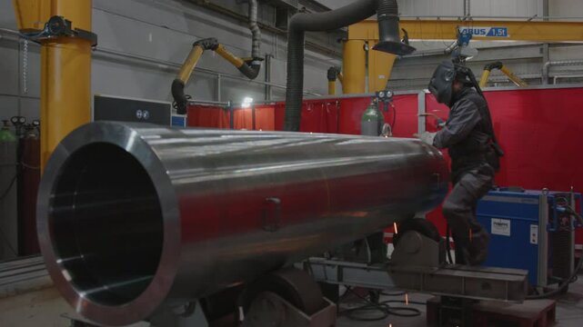 A professional metalworker in a factory welds a large steel pipe with flying sparks and smoke rising from the bright hot welding rod.