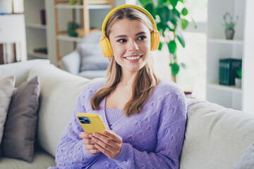 Photo of attractive young woman sit couch headphones hold gadget look empty space dressed purple outfit day light home interior living room