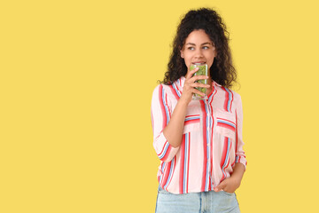 Beautiful young happy African-American woman with glass of tasty kiwi smoothie on yellow background