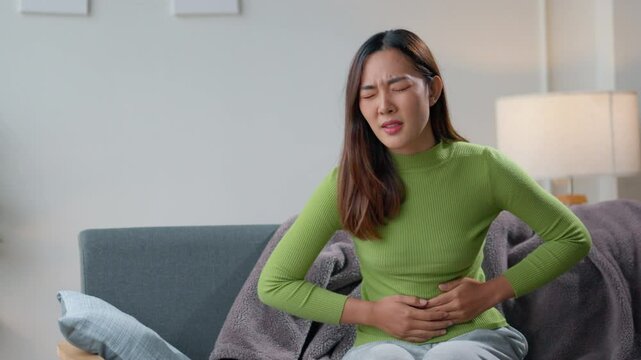 Woman sitting on a sofa, holding her stomach and wincing in pain, indicates a health related issue. Discomfort and concern are evident as she struggles with abdominal pain