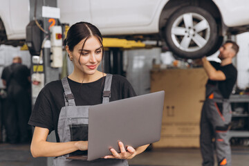 Worker of autoservice, beautiful young woman holding laptop in hands and looking at camera, smiling. Mechanics fixing, repairing automobile lifted on bridge behind.
