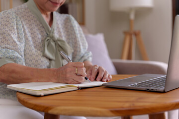 Senior woman sitting at a wooden table with a laptop and taking notes on a notebook
