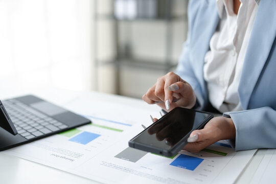 Businesswoman is using a smartphone to analyze financial data, with charts and graphs spread out on the desk in front of her