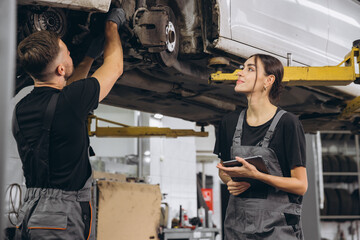 Young expert female inspects repair checklist with automotive mechanic worker partner, quality suspension technician team at fix garage. Vehicle maintenance service works industry occupation job.