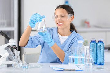 Fototapeta premium Young female scientist in safety goggles and gloves holding erlenmeyer flask with liquid in laboratory