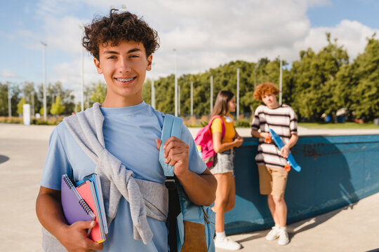Handsome curly teen high school boy guy with braces bag in casual clothes holding netbooks copybooks smiling looking at camera spending time with friends classmates. Students teenagers hanging out