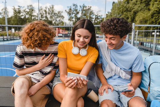 Group of smiling friends teenagers high school pupils college students laughing out having fun watching funny video using gadgets smart phones handing out in sport court yard outside after lessons