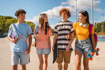 Group of positive teenagers high school pupils friends classmates college students boys and girls with bags skateboards walking in urban street hanging out together talking speaking with each others