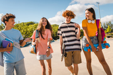 Group of positive teenagers high school pupils friends classmates college students boys and girls with bags holding books skateboards hanging laughing out walking along urban street park