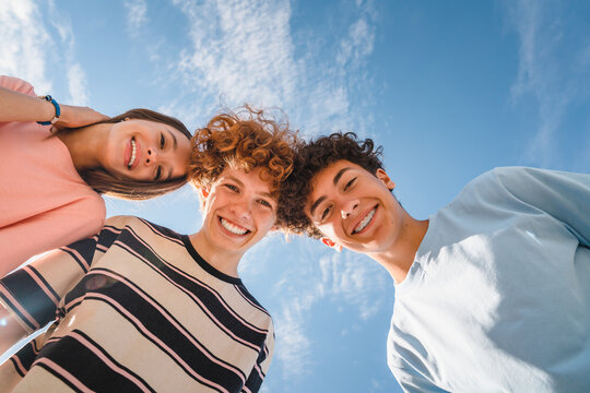 Low angle view of funny smiling teenagers standing and looking down at camera. Group of cheerful friends in casual clothes having fun together outdoors. Happy laughing boys and girls.