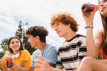 Smiling group of teenagers classmates students school pupils friends boys girls meeting talking have fun break drinking soft drinks fresh juice spend time in skate park sitting on ramp bench outside